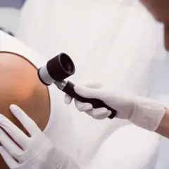 Dermatolog w białej rękawiczce bada skórę pacjenta dermatoskopem.